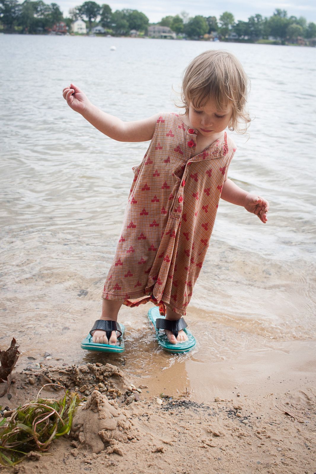 A girl on the edge of a beach, facing the camera, wearing flip flop shoes which are too large for her