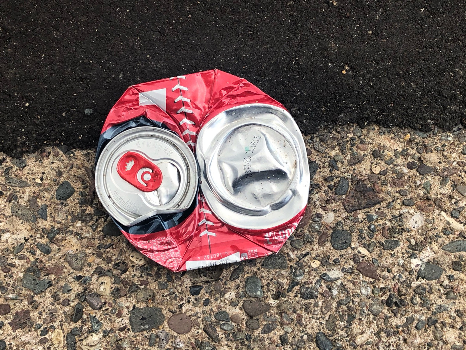 A crushed red and silver aluminum can, sitting on the road right on like line between black asphault and beige curb concrete